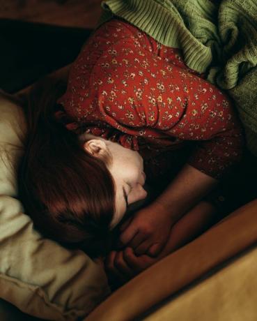 ar zaļu segu apsegtas sarkanmatainas sievietes portrets, kas guļ uz dzeltenā dīvāna un guļ rudens vai ziemas noskaņās, silta, mājīga atmosfēra dzerot rīta kafiju
