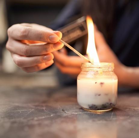 apgriezt anonīmu sievieti ar degošu sērkociņu aizdedzinošu aromātisku sveci stikla burkā, kas novietota uz marmora galda mājās