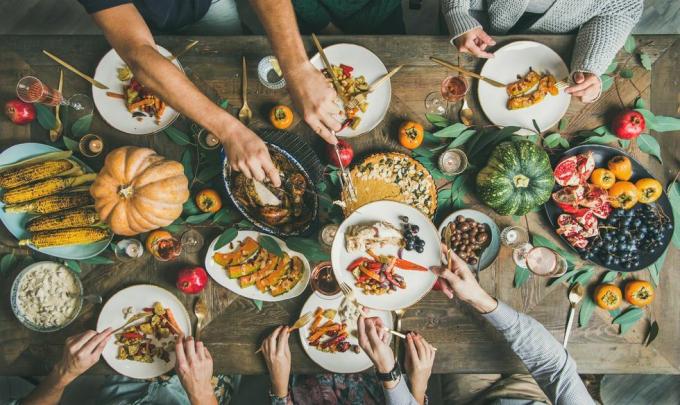 Pateicības dienas galds ar tītaru, ķirbju pīrāgu, grauzdētiem sezonas dārzeņiem un augļiem un cilvēkiem, kas pilda šķīvjus un ēd