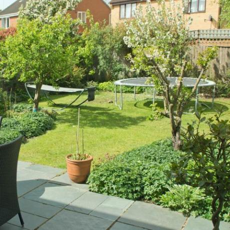 aizmugurējais dārzs angļu piepilsētas īpašumā ar krēsliem un galdu uz šīfera terases un batutu un šūpuļtīklu uz zāliena fonā