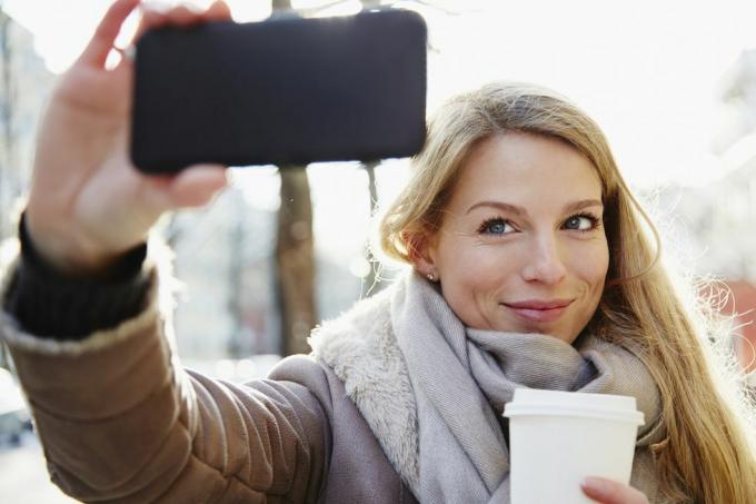 skaista sieviete, kas ziemā staigā pa pilsētas ielām, uzņem selfiju un tur kafiju, lai dotos, aizmugurgaismota