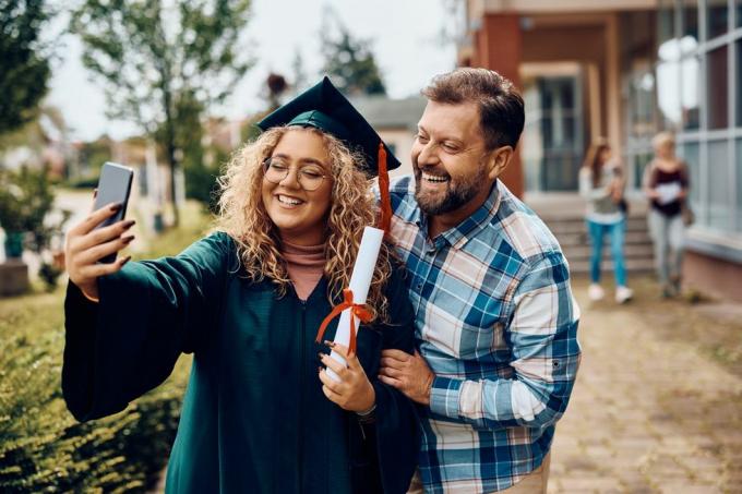 laimīga universitātes absolvente un viņas tēvs izklaidējas, uzņemot selfiju ar viedtālruni