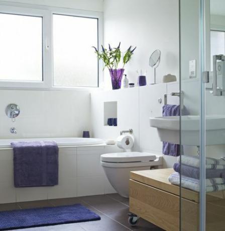 Spilgti apgaismota vannas istaba ar purpura dvieli vannas pusē un salocītus dvieļus pie dušas